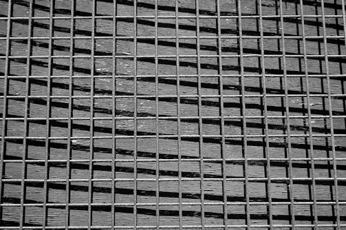 A black and white grid pattern composed of horizontal and vertical lines intersecting to form squares. The grid appears to be made from metal or wire with visible texture and shadows casting diagonal lines across the surface.