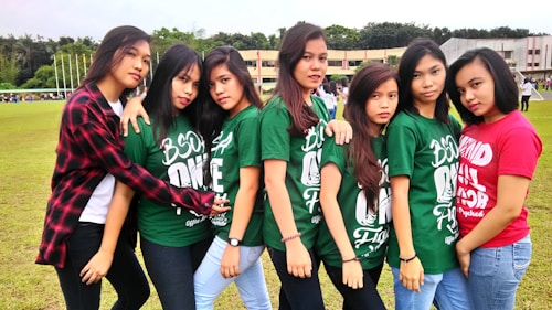 A group of seven young women standing closely together on a grassy field. They are wearing casual clothing, with six of them in matching green shirts and one in a red shirt. The background shows a few buildings and trees, with more people in the distance.