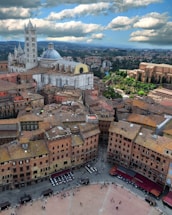 An aerial view of a historic European city featuring a large cathedral with a prominent dome and striped tower. The buildings have terracotta roofs and the city is surrounded by lush greenery. In the foreground is a wide, open plaza with people gathered and outdoor seating visible, suggesting a vibrant communal space.