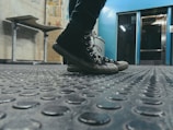 Black sneakers on a textured rubber floor in an industrial setting, with a metallic blue wall in the background. A bench and posters are visible on the wall to the left.