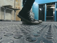 Black sneakers on a textured rubber floor in an industrial setting, with a metallic blue wall in the background. A bench and posters are visible on the wall to the left.