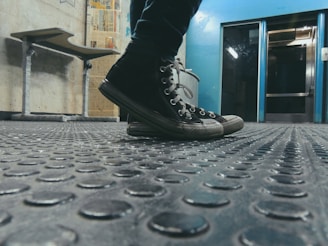Black sneakers on a textured rubber floor in an industrial setting, with a metallic blue wall in the background. A bench and posters are visible on the wall to the left.