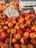 A pile of ripe nectarines is displayed in a wooden crate at a market. A card with the number '60' written on it is prominently placed among the fruit. A digital scale is visible in the background.