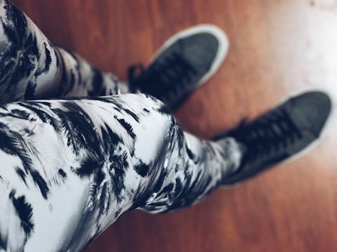 A person is wearing black and white patterned leggings and dark high-top sneakers. The background features a wooden floor.