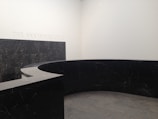 A modern, minimalist interior space with a large, curved black marble wall. The marble has subtle veining and the floor appears to be made of large gray tiles. The words 'THE ANNENBERG COURT' are inscribed on the white wall, suggesting this is part of a building's entry or lobby area.