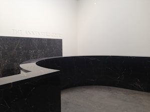 A modern, minimalist interior space with a large, curved black marble wall. The marble has subtle veining and the floor appears to be made of large gray tiles. The words 'THE ANNENBERG COURT' are inscribed on the white wall, suggesting this is part of a building's entry or lobby area.