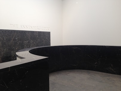A modern, minimalist interior space with a large, curved black marble wall. The marble has subtle veining and the floor appears to be made of large gray tiles. The words 'THE ANNENBERG COURT' are inscribed on the white wall, suggesting this is part of a building's entry or lobby area.