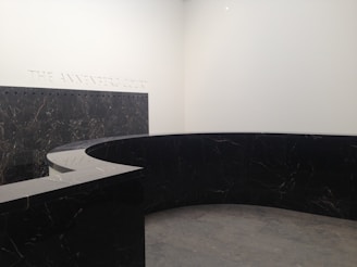 A modern, minimalist interior space with a large, curved black marble wall. The marble has subtle veining and the floor appears to be made of large gray tiles. The words 'THE ANNENBERG COURT' are inscribed on the white wall, suggesting this is part of a building's entry or lobby area.