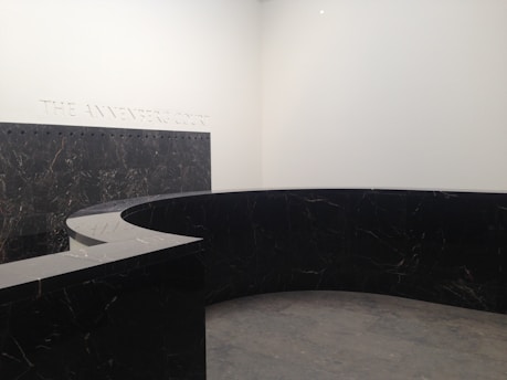 A modern, minimalist interior space with a large, curved black marble wall. The marble has subtle veining and the floor appears to be made of large gray tiles. The words 'THE ANNENBERG COURT' are inscribed on the white wall, suggesting this is part of a building's entry or lobby area.