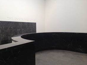 A modern, minimalist interior space with a large, curved black marble wall. The marble has subtle veining and the floor appears to be made of large gray tiles. The words 'THE ANNENBERG COURT' are inscribed on the white wall, suggesting this is part of a building's entry or lobby area.