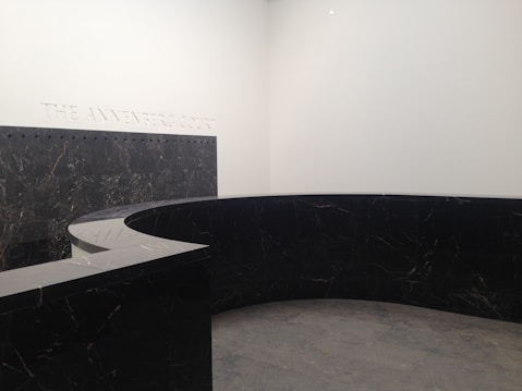 A modern, minimalist interior space with a large, curved black marble wall. The marble has subtle veining and the floor appears to be made of large gray tiles. The words 'THE ANNENBERG COURT' are inscribed on the white wall, suggesting this is part of a building's entry or lobby area.