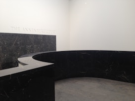 A modern, minimalist interior space with a large, curved black marble wall. The marble has subtle veining and the floor appears to be made of large gray tiles. The words 'THE ANNENBERG COURT' are inscribed on the white wall, suggesting this is part of a building's entry or lobby area.