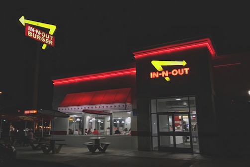 A fast-food restaurant with bright red neon signage and arrows. The building is illuminated in the night with people visible inside through large windows. The exterior has a clean and modern look, with outdoor seating and a distinctive roof design.