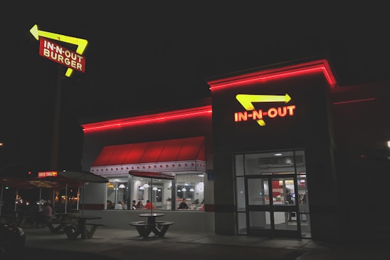 A fast-food restaurant with bright red neon signage and arrows. The building is illuminated in the night with people visible inside through large windows. The exterior has a clean and modern look, with outdoor seating and a distinctive roof design.