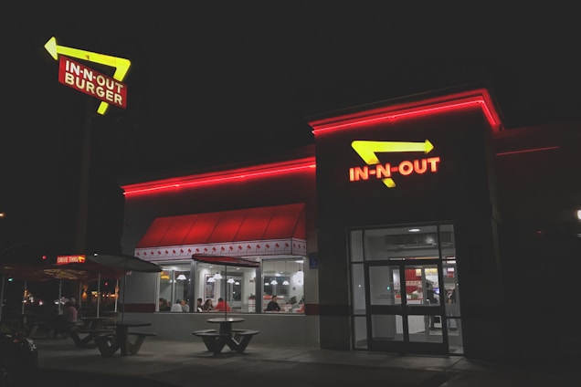 A fast-food restaurant with bright red neon signage and arrows. The building is illuminated in the night with people visible inside through large windows. The exterior has a clean and modern look, with outdoor seating and a distinctive roof design.