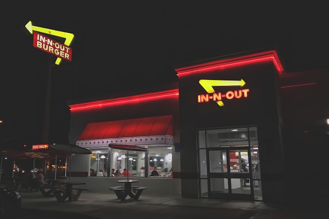 A fast-food restaurant with bright red neon signage and arrows. The building is illuminated in the night with people visible inside through large windows. The exterior has a clean and modern look, with outdoor seating and a distinctive roof design.