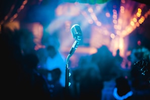 A vintage microphone stands prominently in focus with a blurred background of colorful bokeh lights, suggesting a vibrant and energetic atmosphere. The soft, multicolored lighting creates a dreamy and artistic effect.