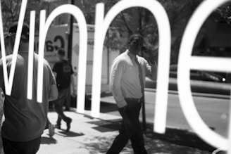 Reflections in a glass window create an abstract scene. Several people are partially visible, with one person in a white shirt and tie standing out. The words on the glass appear large and distorted, encompassing the scene. Trees and a parked truck are also visible in the background, contributing to the urban setting.