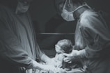 Two medical professionals wearing scrubs, gloves, masks, and goggles are holding and examining a newborn baby. The setting appears to be an operating room or a hospital delivery room under surgical lighting.