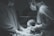 Two medical professionals wearing scrubs, gloves, masks, and goggles are holding and examining a newborn baby. The setting appears to be an operating room or a hospital delivery room under surgical lighting.