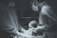 Two medical professionals wearing scrubs, gloves, masks, and goggles are holding and examining a newborn baby. The setting appears to be an operating room or a hospital delivery room under surgical lighting.