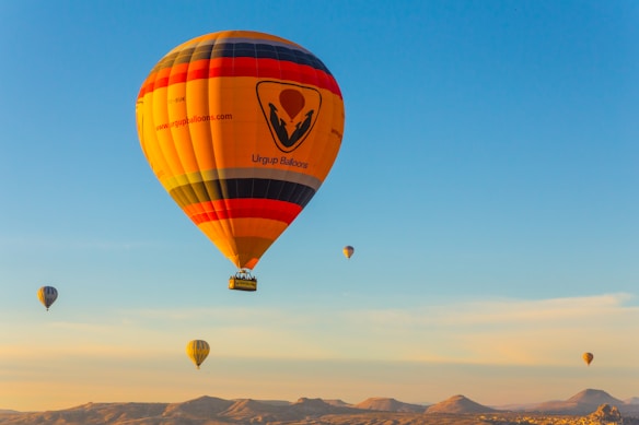 Several hot air balloons float gracefully in the sky over a landscape with rolling hills. The largest balloon is prominently featured with vibrant colors and a logo. The sky is a clear blue, enhancing the visual appeal and sense of peacefulness.