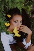A person with curly dark hair is surrounded by lush green foliage and vibrant yellow flowers. The individual has an introspective expression and is wearing a white outfit.