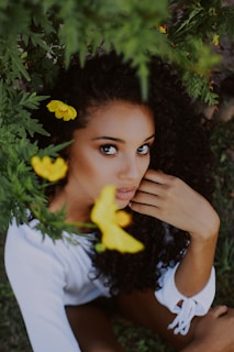 A person with curly dark hair is surrounded by lush green foliage and vibrant yellow flowers. The individual has an introspective expression and is wearing a white outfit.