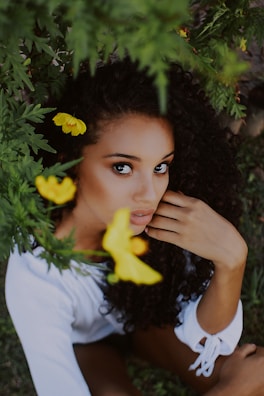 A person with curly dark hair is surrounded by lush green foliage and vibrant yellow flowers. The individual has an introspective expression and is wearing a white outfit.