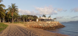 A beautiful beach scene featuring a sandy shoreline, palm trees, a rocky embankment, and a building with a modern design. The sun is low, casting a warm glow on the sand, trees, and rocks. In the background, calm ocean waves gently lap against the shore, and distant mountains are visible on the horizon under a partly cloudy sky.