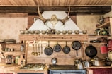 A rustic kitchen interior features a wooden shelf adorned with a row of ornate teacups and a mounted antler above. Below, several hanging frying pans and cooking utensils are neatly arranged. The countertop displays a gas stove, and various pots and items, including a kettle, are visible. The decor includes a traditional theme with exposed wooden beams and textured wallpaper.