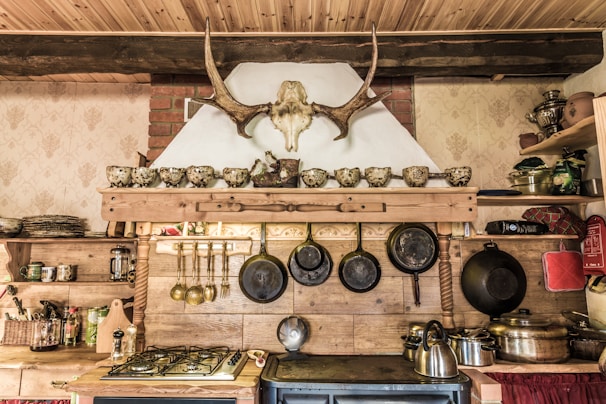 A rustic kitchen interior features a wooden shelf adorned with a row of ornate teacups and a mounted antler above. Below, several hanging frying pans and cooking utensils are neatly arranged. The countertop displays a gas stove, and various pots and items, including a kettle, are visible. The decor includes a traditional theme with exposed wooden beams and textured wallpaper.