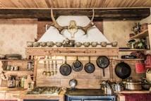 A rustic kitchen interior features a wooden shelf adorned with a row of ornate teacups and a mounted antler above. Below, several hanging frying pans and cooking utensils are neatly arranged. The countertop displays a gas stove, and various pots and items, including a kettle, are visible. The decor includes a traditional theme with exposed wooden beams and textured wallpaper.