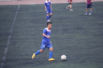 A soccer player in a blue uniform and yellow shoes is running on a dark green field while controlling a white soccer ball. Other players in similar uniforms and a red and blue striped uniform are visible in the background.
