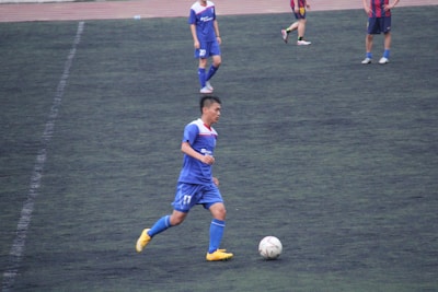 A soccer player in a blue uniform and yellow shoes is running on a dark green field while controlling a white soccer ball. Other players in similar uniforms and a red and blue striped uniform are visible in the background.