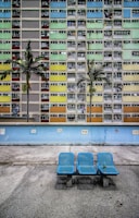 A colorful residential building with a grid of windows displaying various colors, including pastel blue, green, yellow, and peach. Two palm trees stand in front of the building, and there is a light blue wall separating the building from the foreground. In the foreground, three blue seats, with one holding a green bottle, are placed on a gray, speckled surface.