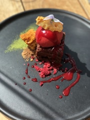 A beautifully plated dessert featuring a red scoop resembling a sorbet on top of a chocolate brownie. Accompanied by a piece of honeycomb and garnished with edible flowers. There’s a green powder, possibly matcha, and a drizzle of red sauce with crumbled red bits, all served on a black matte plate.