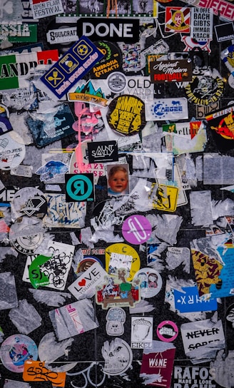 A chaotic collage of various stickers and posters overlapping each other on a dark surface. The stickers feature different designs, logos, and text, with a mixture of colors and styles. Some are partially torn or obscured, creating a layered appearance.