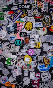 A chaotic collage of various stickers and posters overlapping each other on a dark surface. The stickers feature different designs, logos, and text, with a mixture of colors and styles. Some are partially torn or obscured, creating a layered appearance.