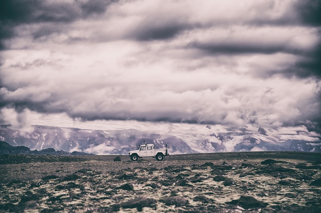 A rugged landscape under a dramatic cloudy sky with a white off-road vehicle parked on a rocky terrain. Snow-covered mountains form the distant background, adding a sense of isolation and adventure.