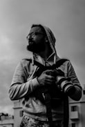 A black and white photograph of a person wearing glasses, a hoodie, and carrying a camera with a large lens. The person appears to be outdoors, with buildings faintly visible in the background. The individual is looking to the side with a focused expression.
