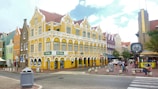 A vibrant city street scene with a yellow colonial-style building featuring white trim and arched windows. People are casually walking and sitting around the area. A large clock stands on a pole near the corner of the street, and pedestrian crosswalks are visible. The sky is partly cloudy and other colorful buildings can be seen in the background.