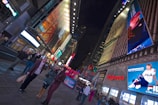 A bustling city street scene with large, brightly lit billboards displaying various advertisements. People are walking and interacting on the sidewalk. The atmosphere is vibrant and lively, typical of a busy urban area at night.