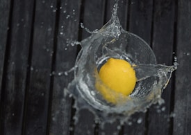 A lemon is splashing into a clear glass filled with water, creating dynamic water splashes around it. The background consists of dark wooden planks that provide a contrast to the vibrant yellow of the lemon.
