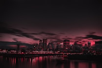 A cityscape at dusk or nighttime with buildings silhouetted against a dark sky featuring dramatic red and black hues. The reflection of city lights creates a mirrored effect on the water in the foreground.