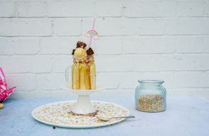 A glass mug with a creamy milkshake topped with a scoop of ice cream, chocolate pieces, and colorful sprinkles is positioned on a pedestal. It features a straw with a decorative rainbow. The background includes a white brick wall and a jar filled with more sprinkles is nearby.