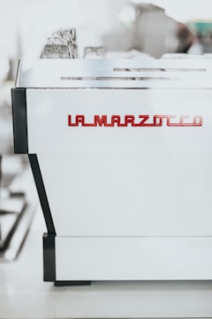 A professional espresso machine with a sleek, modern design. The machine is prominently labeled with 'La Marzocco' in red, positioned against a shiny white surface with black accents.