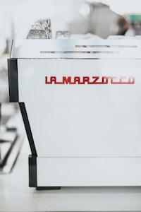 A professional espresso machine with a sleek, modern design. The machine is prominently labeled with 'La Marzocco' in red, positioned against a shiny white surface with black accents.