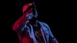 A performer wearing a light-colored jacket and a cap holds a microphone close to their mouth against a dark background. The scene is dimly lit, creating contrasting shades with red and blue lighting casting dramatic shadows.