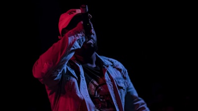 A performer wearing a light-colored jacket and a cap holds a microphone close to their mouth against a dark background. The scene is dimly lit, creating contrasting shades with red and blue lighting casting dramatic shadows.